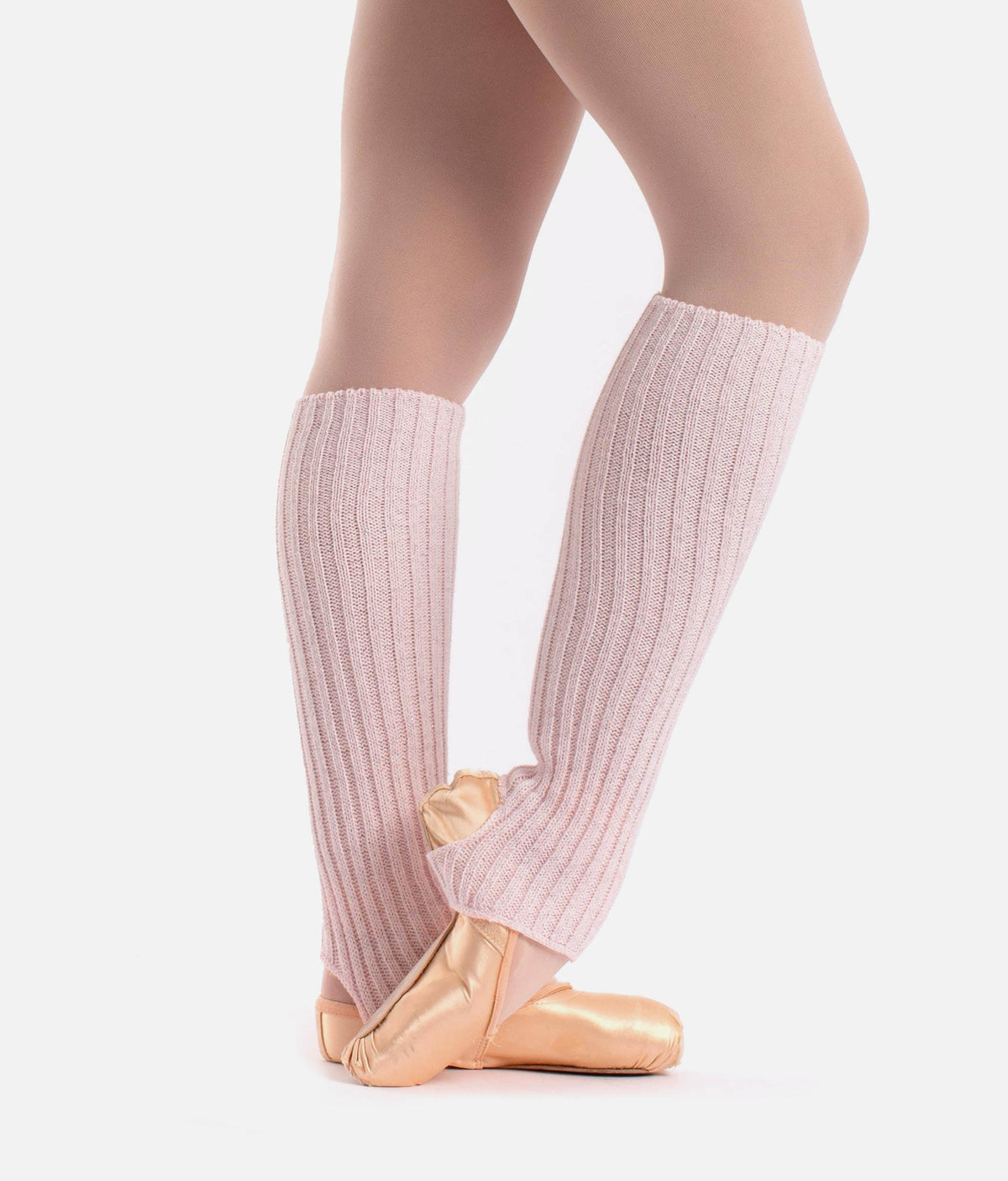 A close-up of a dancer's legs wearing light pink ribbed knee-high leg warmers over nude tights and metallic gold ballet slippers, set against a plain light gray background.