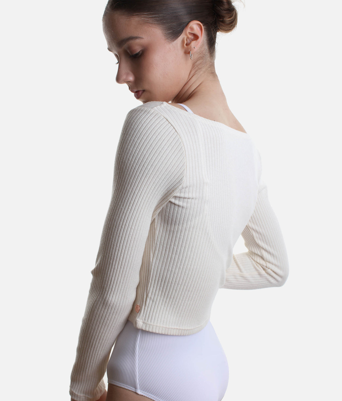 A woman shown from the back wearing a ribbed long-sleeve crop top in off-white, paired with white high-waisted bottoms, against a plain light gray studio background.