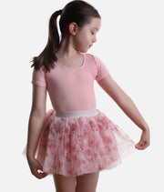 A young girl with a ponytail wearing a pink short-sleeve leotard and a pink and white patterned tutu skirt, posing against a light gray background.