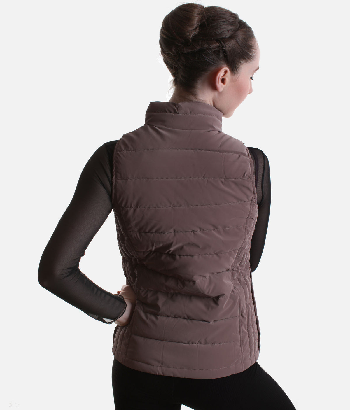 A woman shown from the back wearing a taupe-brown quilted puffer vest over a black long-sleeve top and black leggings, against a light gray studio background.