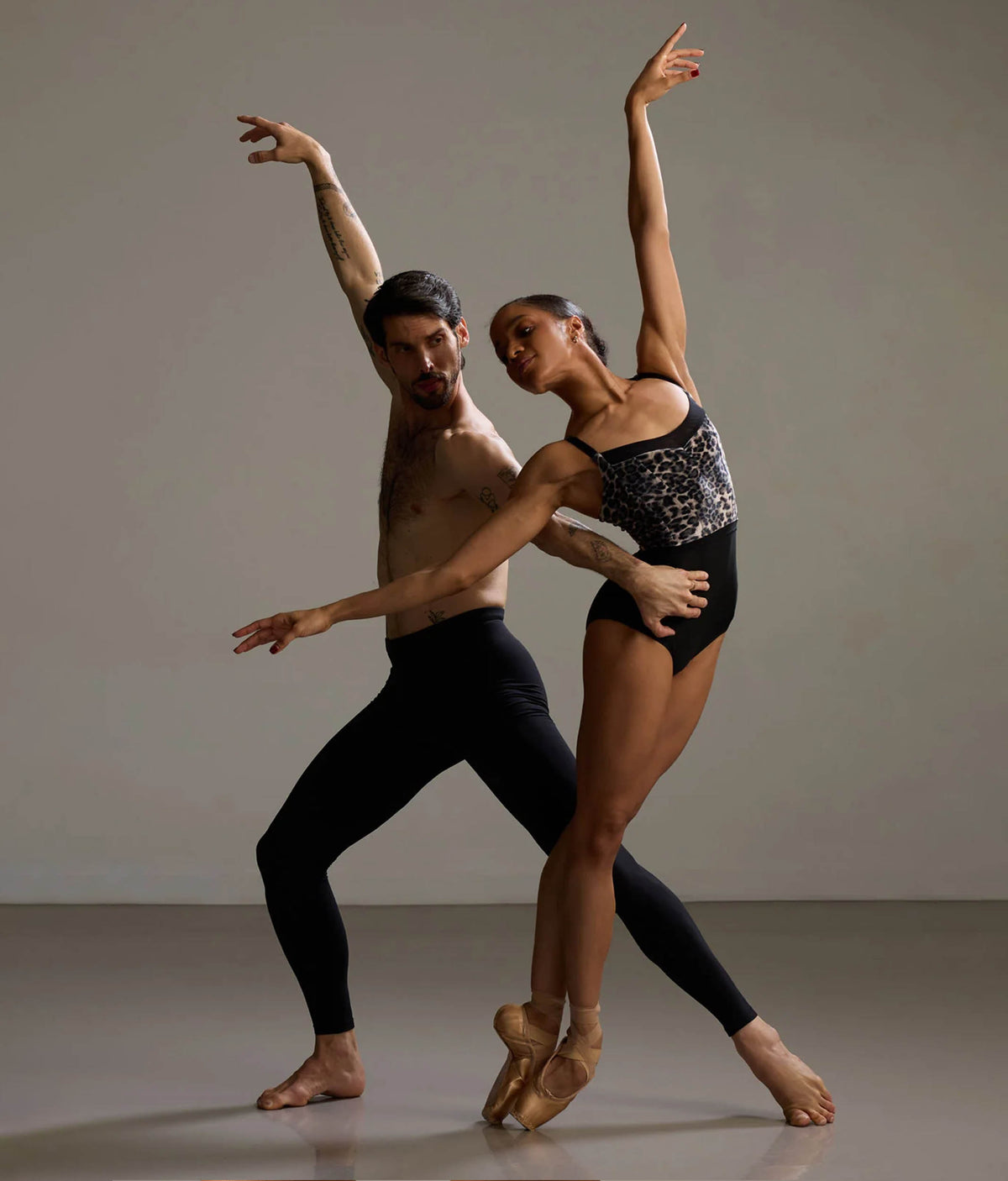 A male and female ballet dancer pose together in a studio. The woman wears a black leotard with a silver patterned bodice and tan pointe shoes, while the man wears black fitted pants and is shirtless.