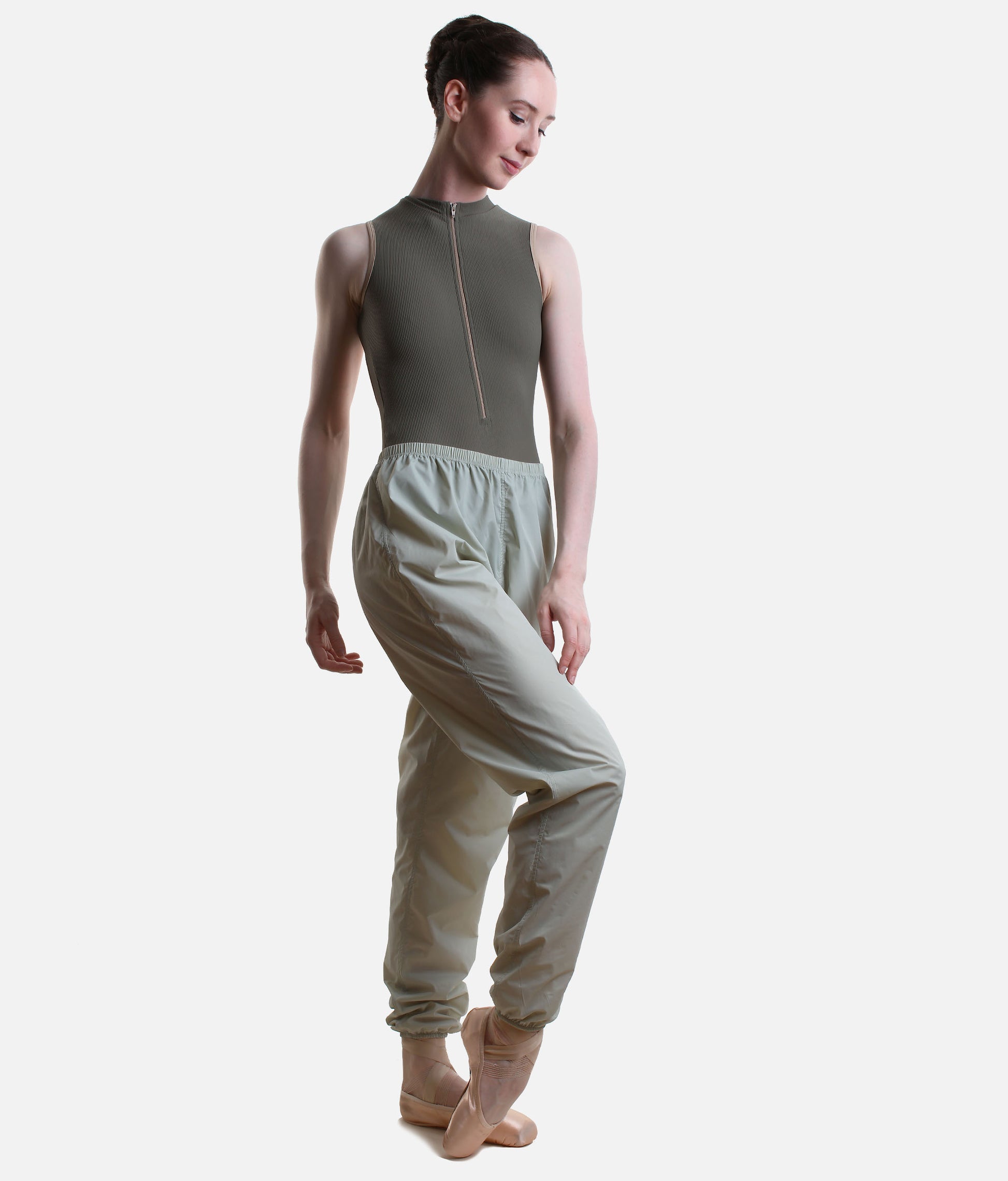 A model wearing a sleeveless olive green dance leotard with a front zipper and matching high-waisted light sage green dance pants, posed against a solid light gray background.