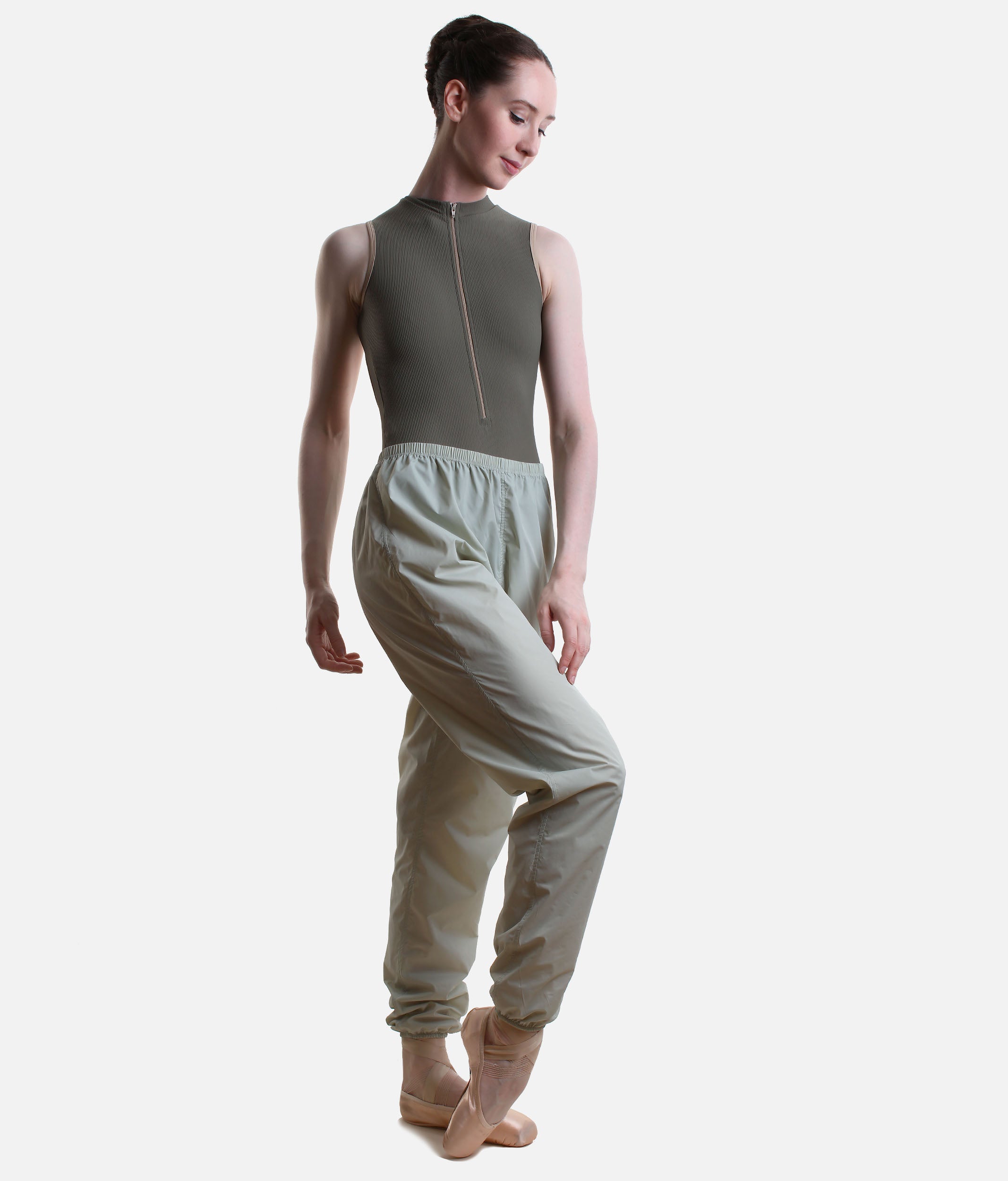 A model wearing a sleeveless olive green dance leotard with a front zipper and matching high-waisted light sage green dance pants, posed against a solid light gray background.
